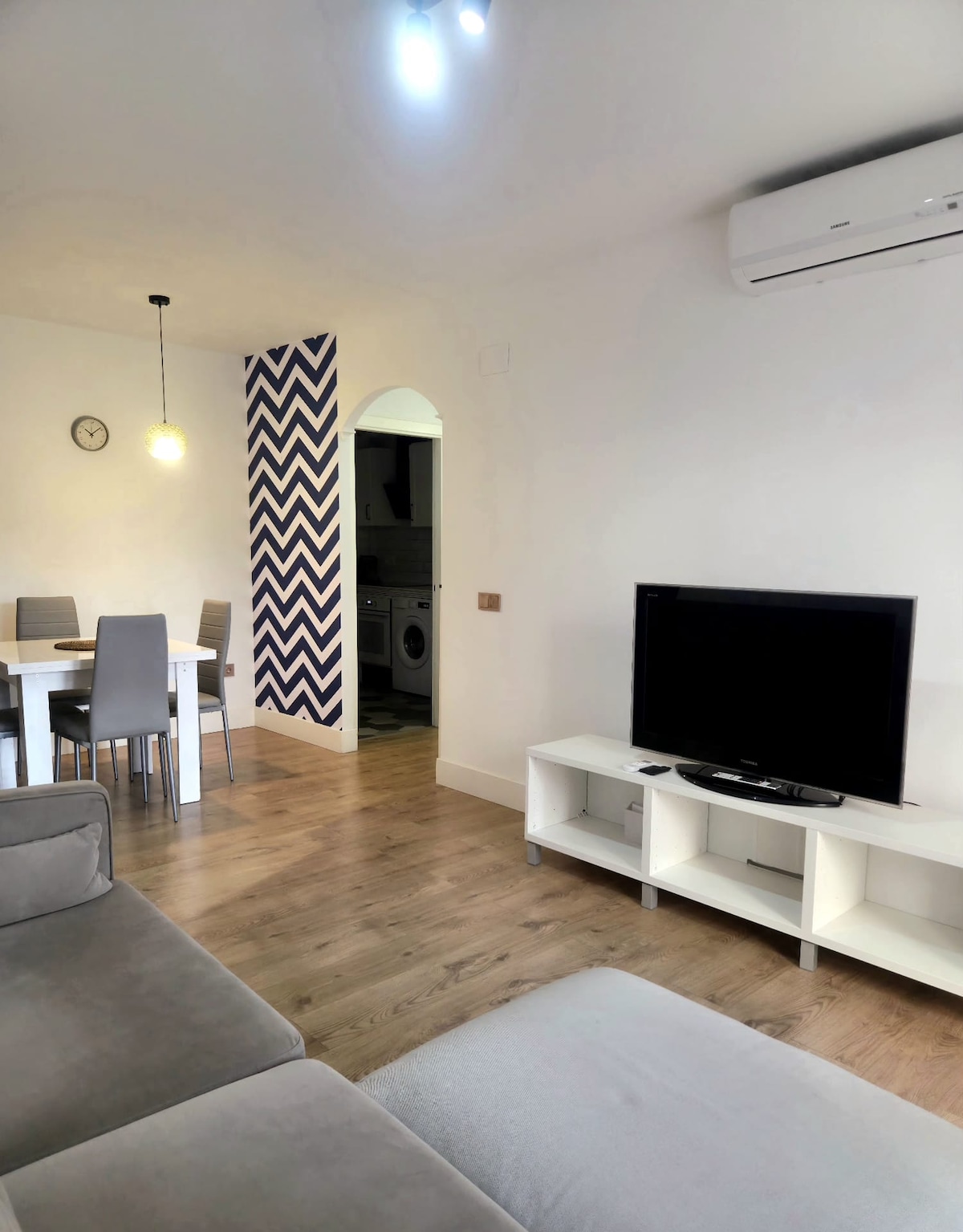 A modern living area features a grey sofa and a flat-screen television on a white media unit. A dining area with a table and four chairs is visible, alongside a wall decorated with a chevron pattern. An air conditioning unit is mounted on the wall, providing comfort.