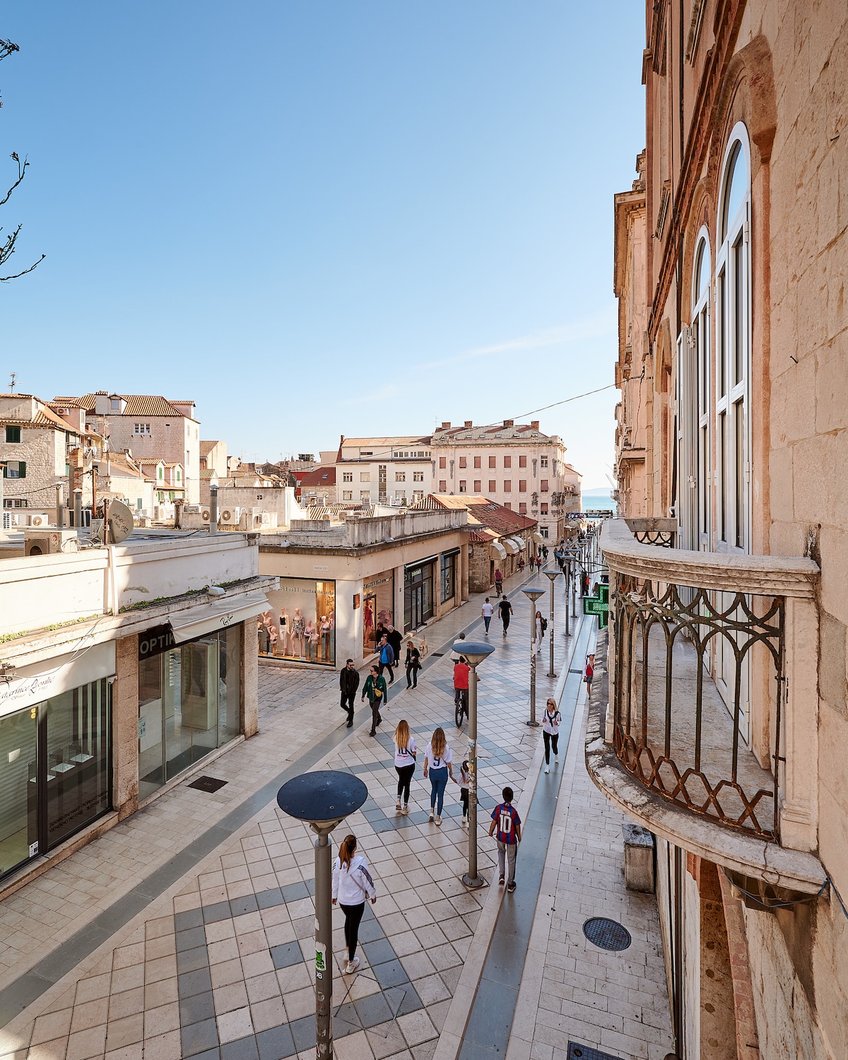 A vibrant street view showcases a bustling pedestrian area lined with charming shops and cafes. The scene is framed by historic buildings, with a clear blue sky above. People can be seen strolling along the walkway, contributing to the lively atmosphere.