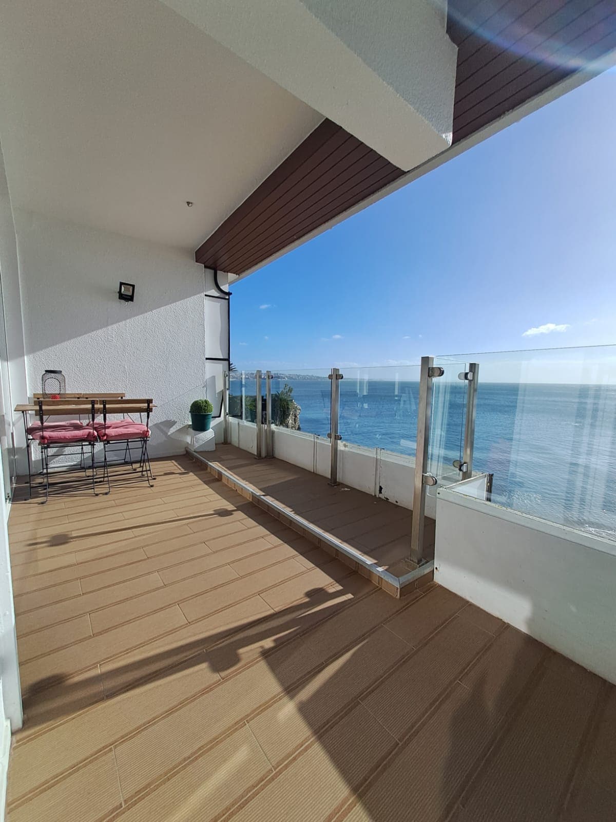 A balcony with floor-to-ceiling glass railings presents an unobstructed view of the ocean. The space includes a small round table and two chairs, inviting relaxation. Sunlight reflects off the water, highlighting the clear sky above.