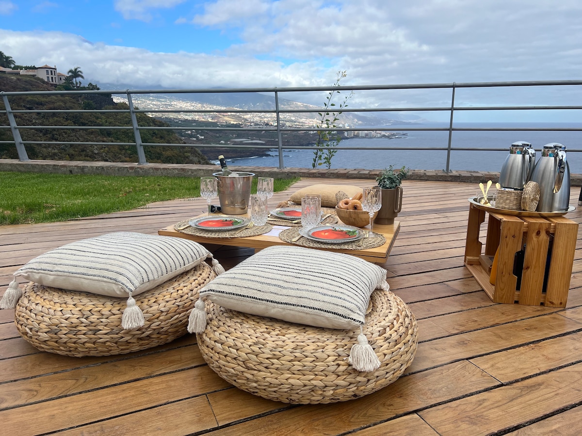 An outdoor seating area is presented on a wooden deck, featuring woven poufs and a low table set with plates and glasses. A scenic view of the coastline and mountains is visible in the background, framed by a railing that enhances the open space.