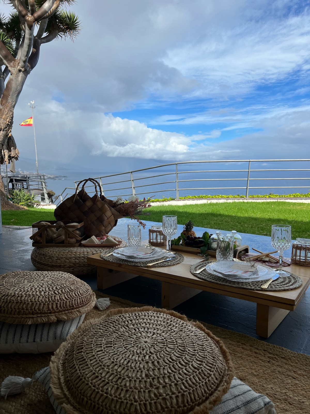 A cozy outdoor dining area features a low wooden table set with glassware and woven placemats. Natural fiber seating surrounds the table. In the background, a lush green lawn meets a panoramic view of the ocean under a partly cloudy sky.