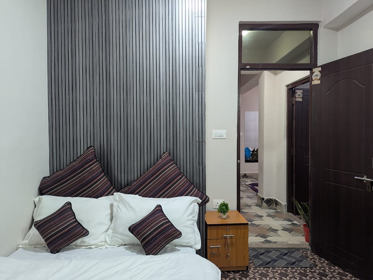 A cozy sleeping area features a spacious bed adorned with decorative cushions. A bedside table with a small plant is positioned beside the bed, while a doorway leads to other areas within the apartment, providing easy access to the surrounding spaces.