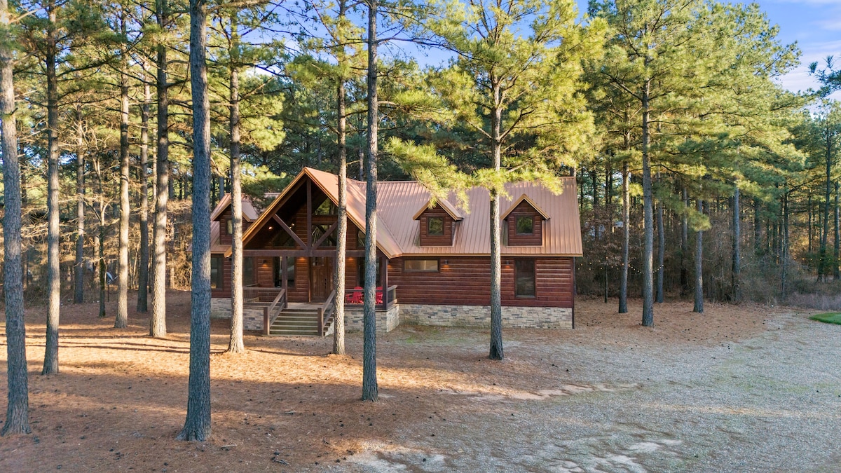 A luxury cabin is set amid tall pine trees, featuring a wooden exterior with a warm-toned roof. The walkway leads to a welcoming entrance, surrounded by a natural, uncluttered landscape covered with a layer of pine needles.