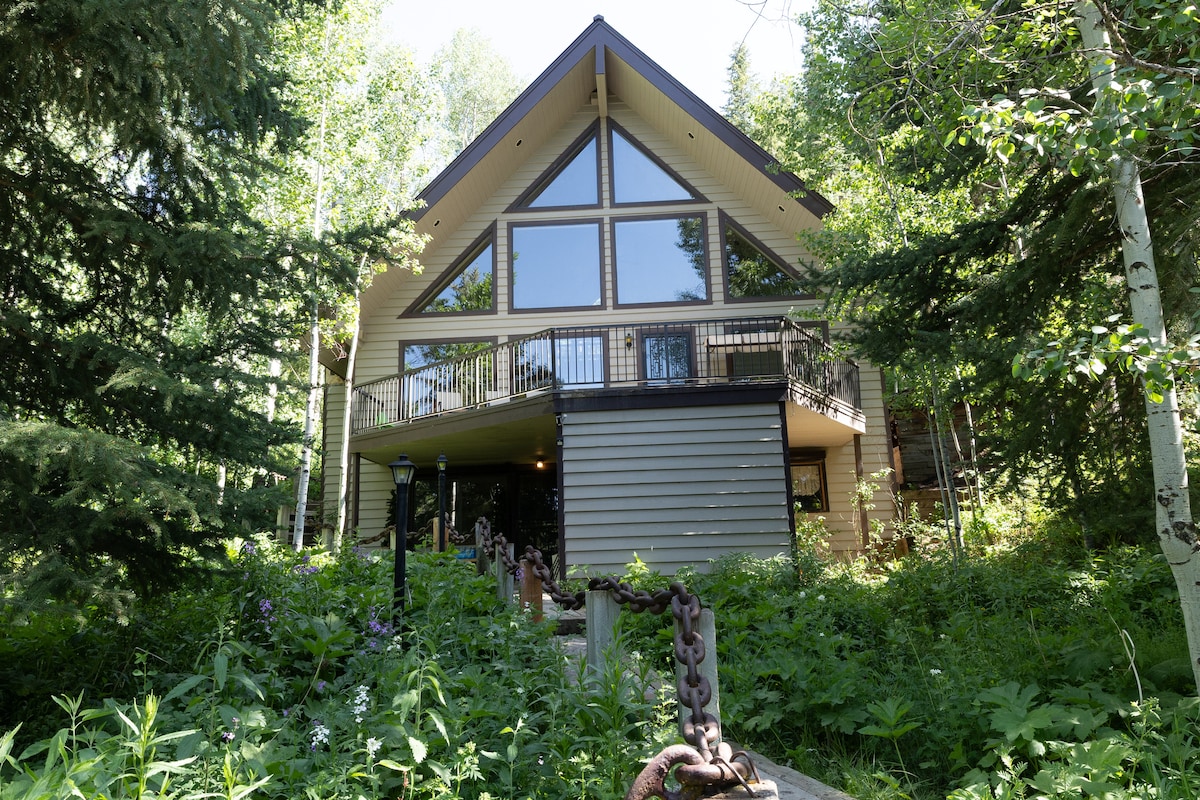The cabin's exterior displays a triangular architecture with large glass windows allowing natural light to illuminate the interior. Surrounding greenery enhances the tranquil setting, while a welcoming entrance features a stone pathway bordered by lush foliage.