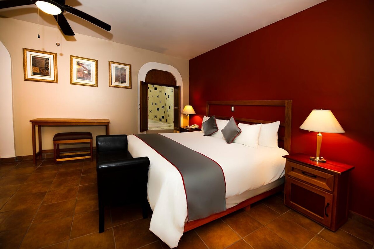 A well-appointed room is highlighted by a spacious bed adorned with neutral-colored linens and accent pillows. Warm red walls contrast with the lighter tones used throughout. A wooden nightstand and lamp complement the furniture, while framed artwork adds character to the inviting space.
