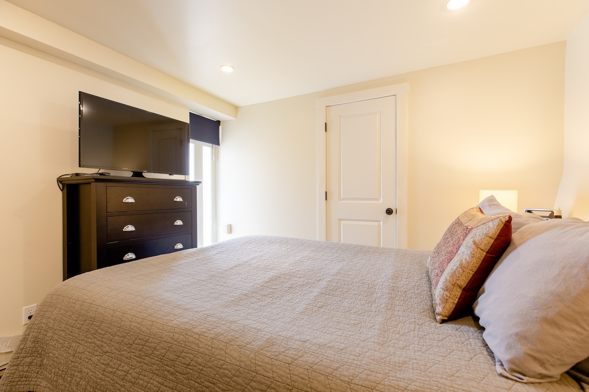 A spacious sleeping area is seen with a queen-sized bed featuring a neutral quilt and decorative pillows. A flat-screen television is mounted on the wall opposite the bed. A door leads to the room, with natural light streaming in through a nearby window adorned with a black-out shade.