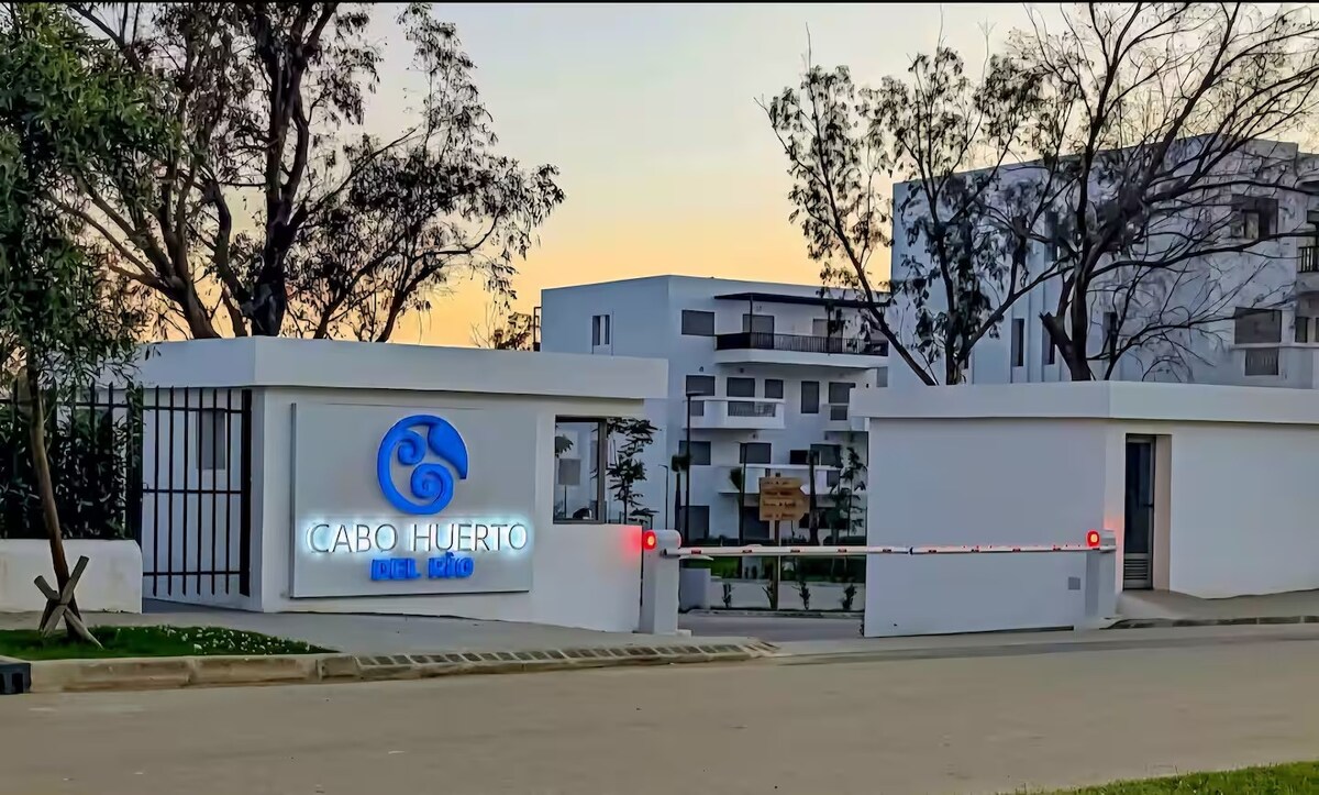 The entrance to the Cabo Huerto complex is marked by a modern sign featuring a blue swirl design. The white-walled structure is set against a soft sunset backdrop, providing a welcoming view of the surrounding greenery and residential buildings.