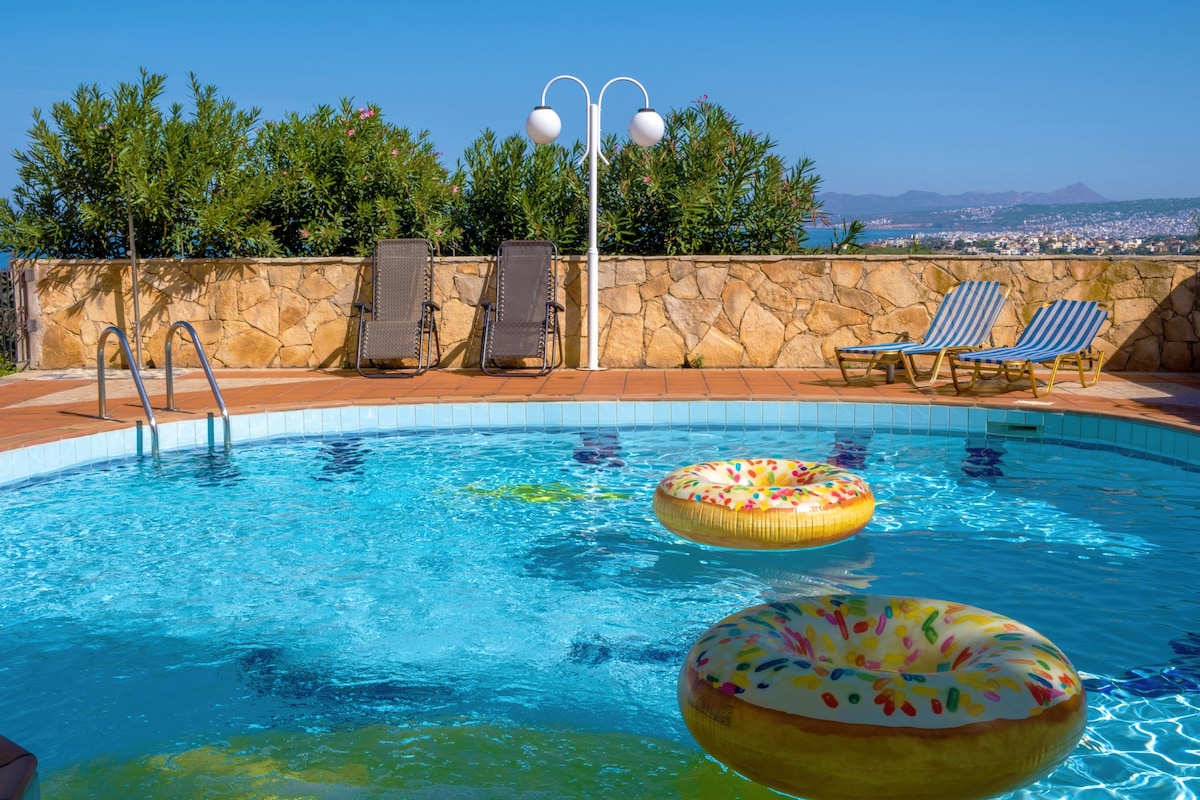 A private pool is showcased, featuring colorful floating rings nestled within the clear blue water. Sun loungers are positioned nearby against a stone wall, complemented by greenery in the background. A soft blue sky adds to the serene beachside atmosphere.