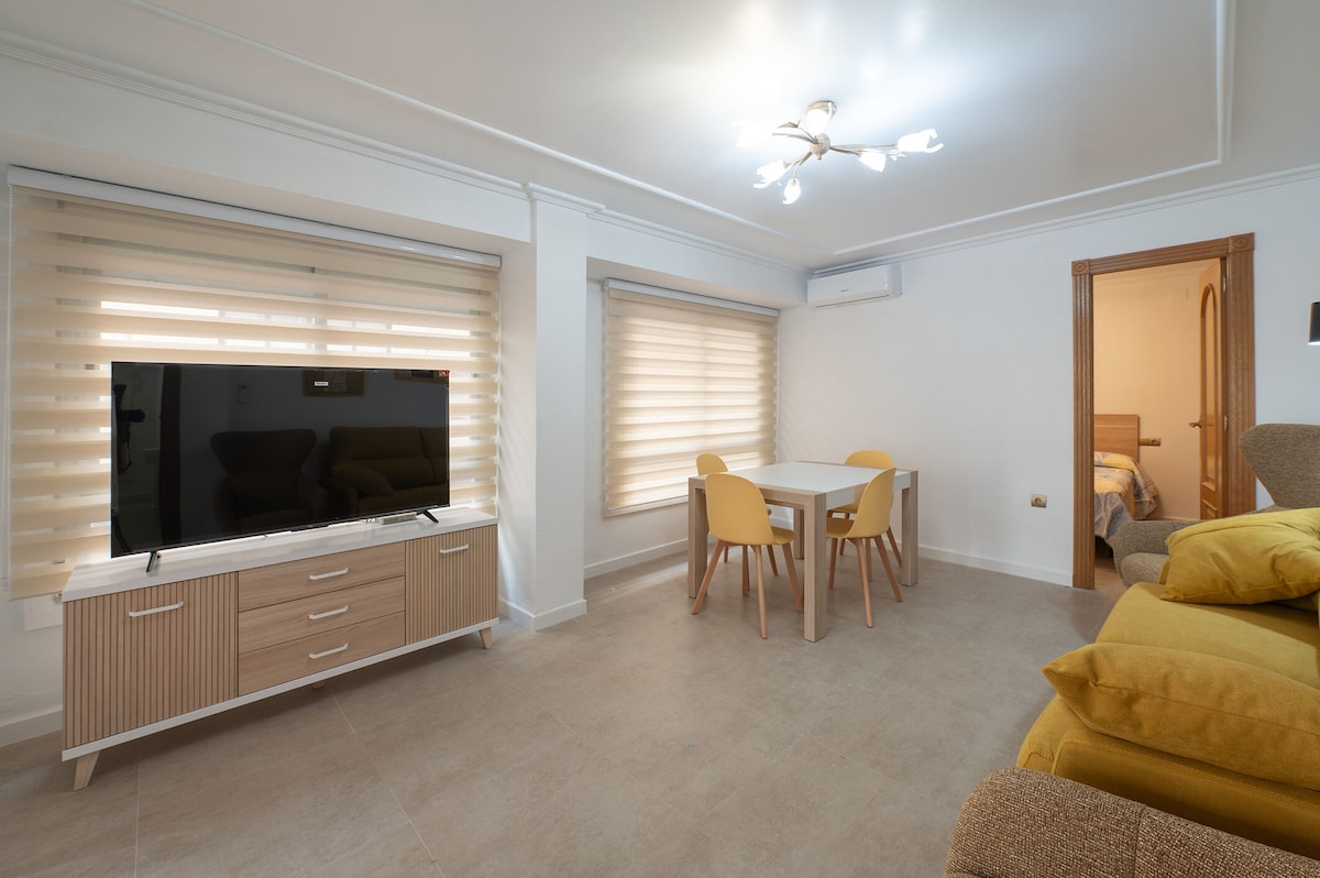 A well-lit living area is presented with a flat-screen TV on a light wood cabinet. A white dining table with four chairs is positioned nearby, while soft yellow cushions are arranged on a comfortable sofa. Window shades diffuse natural light in the inviting space.
