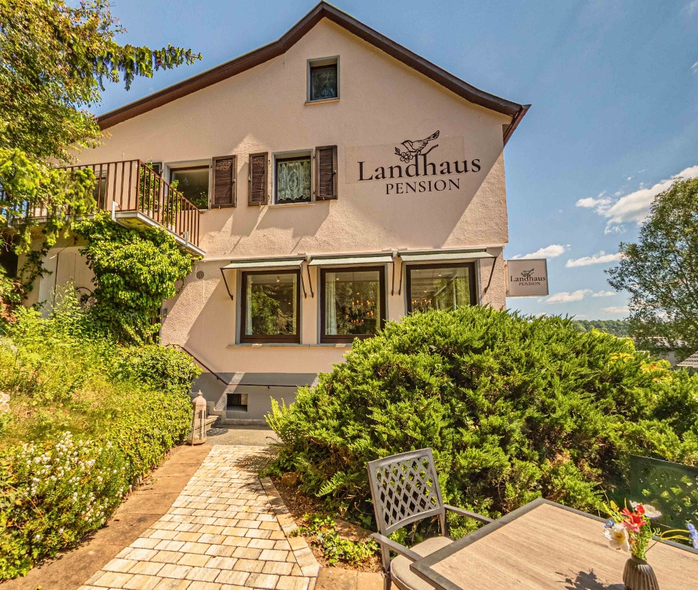 The charming exterior of the guesthouse is highlighted, featuring a light-colored facade and brown shutters. Lush greenery surrounds the entrance, and a stone pathway leads to a seating area with a table and chairs, providing a welcoming view of the property.