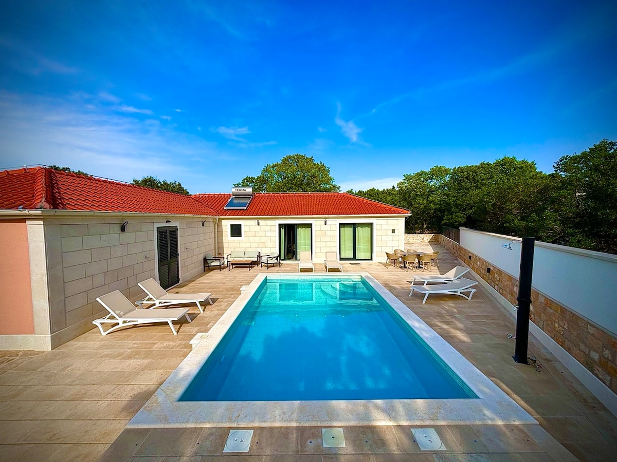 An outdoor area features a private swimming pool surrounded by a tiled terrace. Sun loungers are positioned on one side, while large glass doors lead to the villa. Lush green trees provide a backdrop, and the sky is clear with a gentle blue hue.