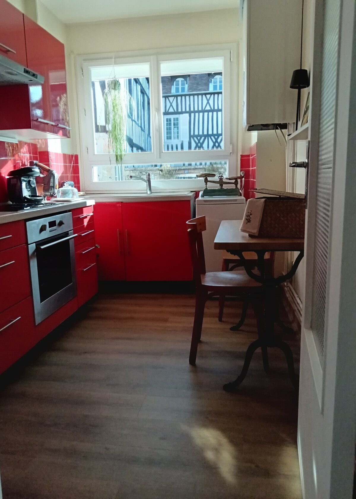 A compact kitchen features red cabinetry and modern appliances. A small table with a chair is positioned next to a window, allowing natural light to illuminate the space. The view includes traditional half-timbered houses, enhancing the kitchen's inviting atmosphere.