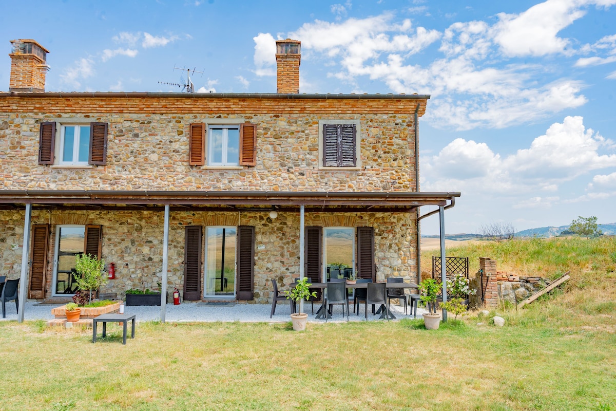 The two-story stone farmhouse features a covered porch with dining furniture, surrounded by a spacious lawn. Large windows allow for natural light, while potted plants add greenery to the outdoor area. Fields extend in the background, complementing the rustic charm of the home.