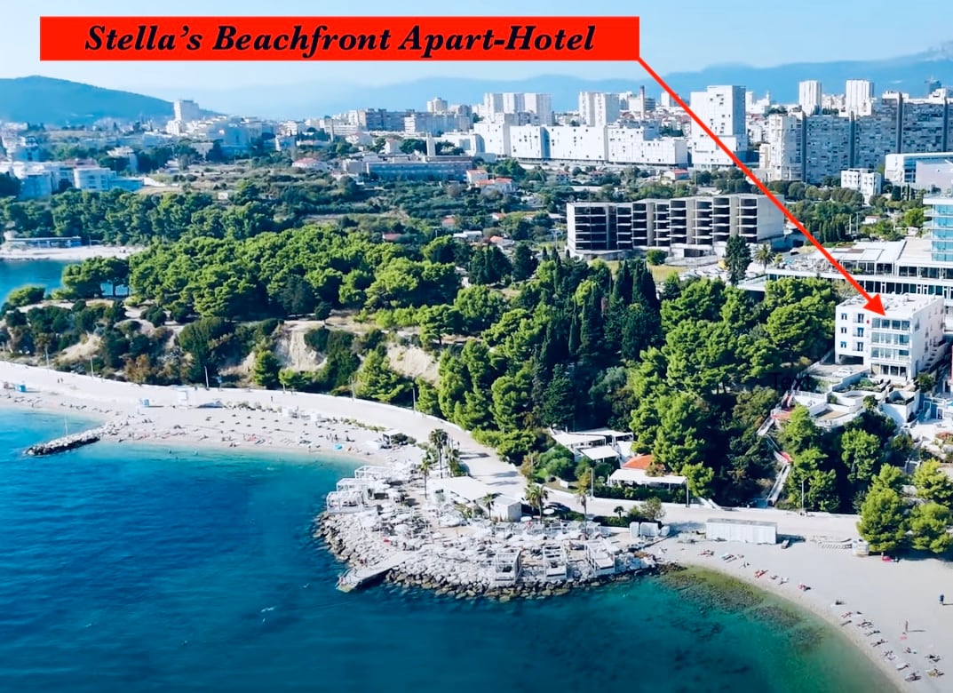 An aerial view showcases Stella's Beachfront Aparthotel, positioned directly on the beachfront. The natural coastline curves gracefully, with sandy beaches and clear waters of the Adriatic Sea. Lush greenery surrounds the property, complemented by modern buildings in the background.