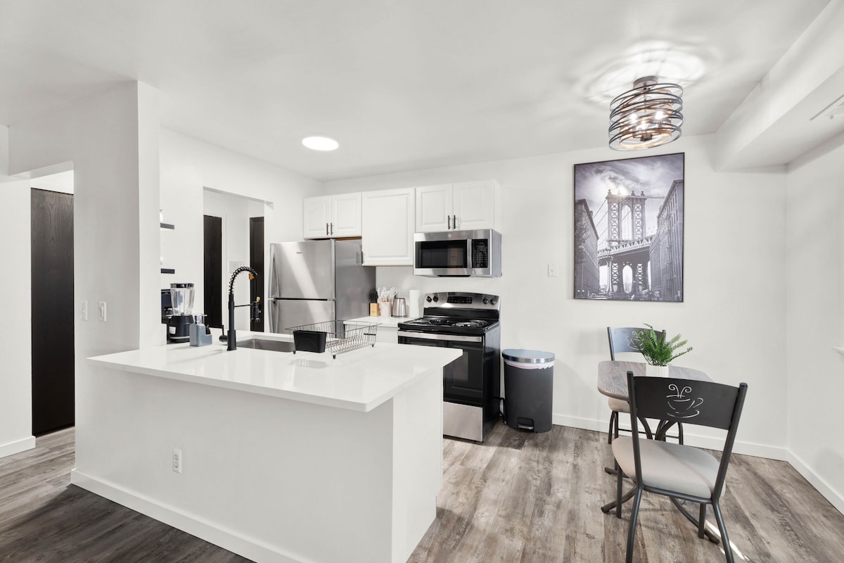 The kitchen features modern appliances, including a stainless steel refrigerator, electric stove, and microwave. A central island with a white countertop provides space for meal preparation, alongside a dining table for two. A decorative wall art piece and a potted plant add visual interest.