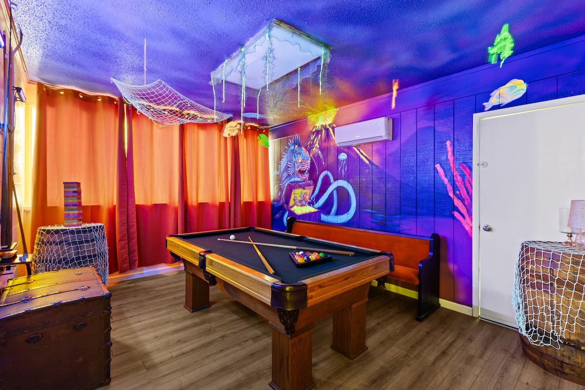 A game room is presented with a vibrant UV-reactive decor, featuring glowing underwater elements. A billiards table occupies the center, complemented by wooden benches. Bright orange curtains allow filtered light, creating a playful atmosphere. Walls are adorned with sea-themed art and decorative nets.