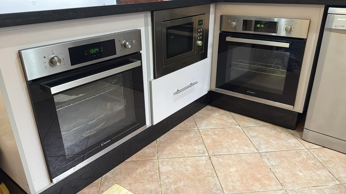 A modern kitchen corner displays three sleek built-in cooking appliances, including two ovens and a microwave. The appliances feature stainless steel finishes with digital controls. A light-colored countertop complements the contemporary look, while tiles on the floor add a subtle texture.