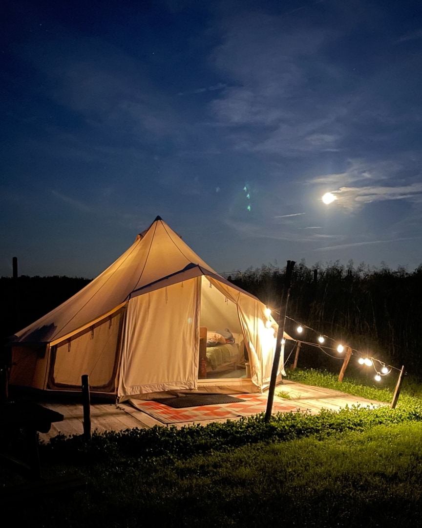 A bell tent is illuminated by soft lighting under a night sky, with a full moon visible in the background. The tent features an open entrance, revealing a glimpse of the interior, while a wooden deck is adorned with a patterned rug, surrounded by lush greenery.