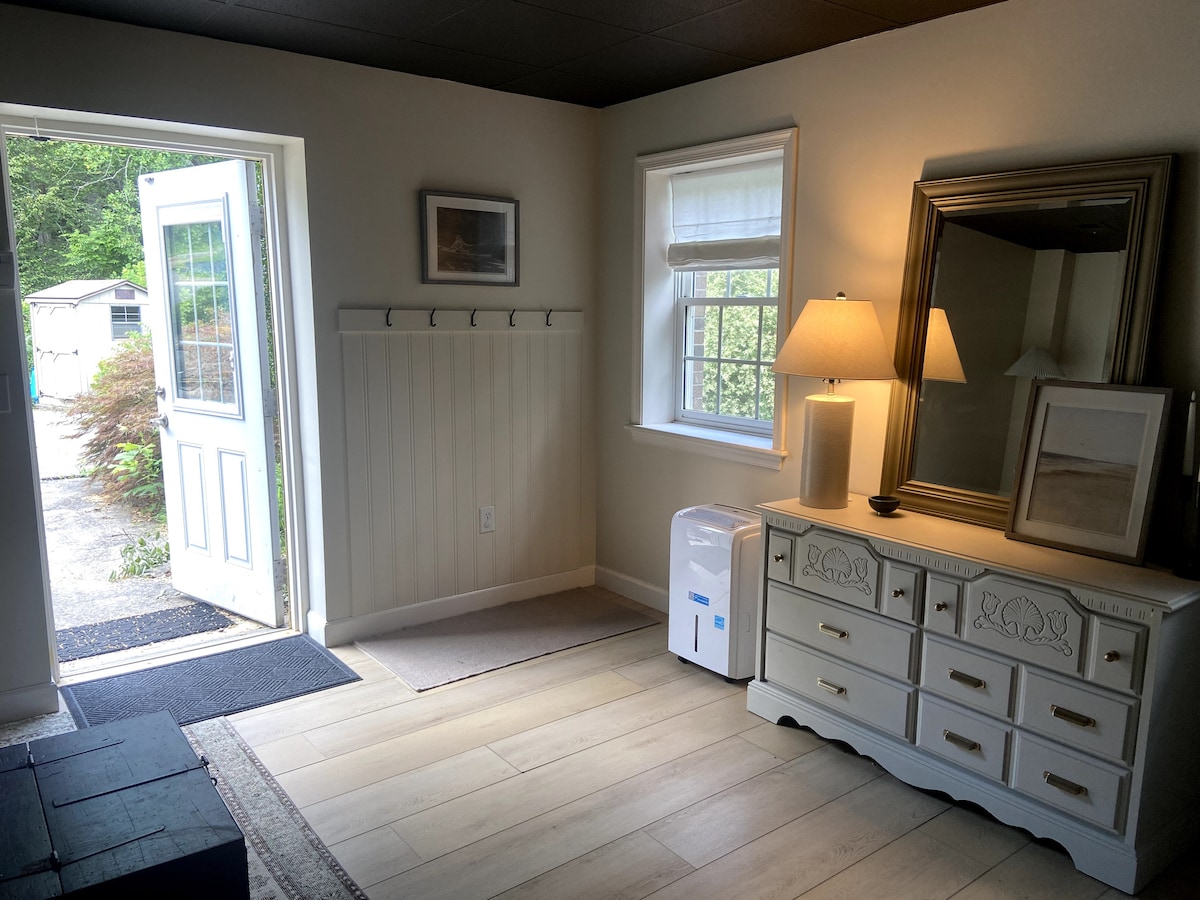 The entrance features a welcoming space with a white door open to the outside. A decorative mirror and a lamp are placed on a white dresser, while a coat rack lines the wall. Natural light illuminates the room, creating a bright ambiance.