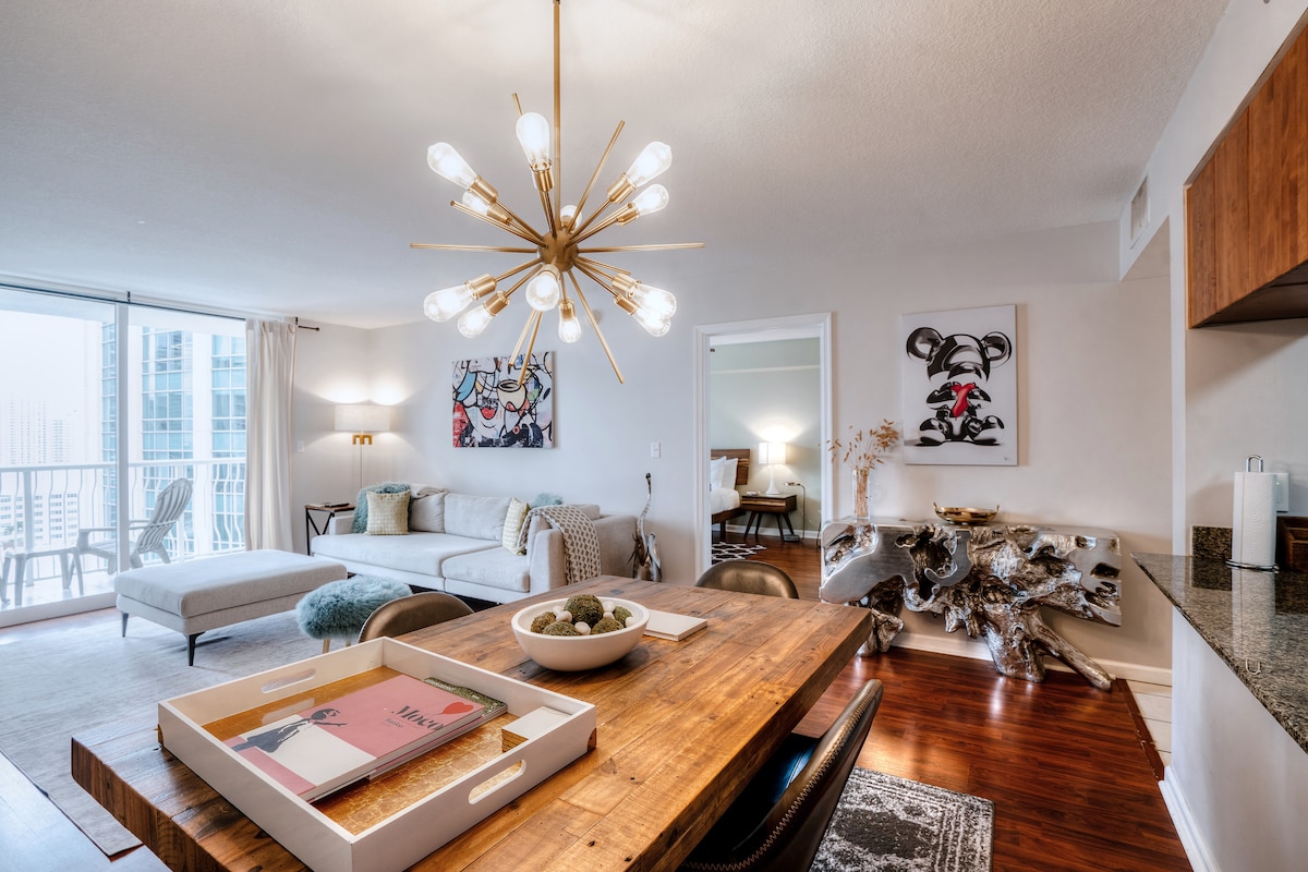 The living area features a modern design with ample natural light flowing through large glass doors leading to a private terrace. A stylish dining table sits adjacent to a fully equipped kitchen, with a decorative console table and unique artwork enhancing the ambiance.