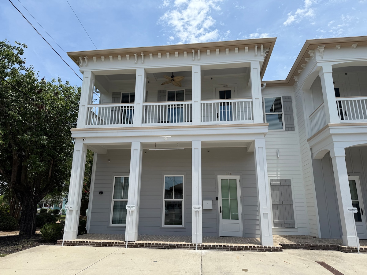 The exterior of a newly renovated first-floor condo showcases a light-colored façade with a welcoming porch. Tall columns support a balcony above, while large windows allow natural light to flood in. Surrounding greenery enhances the pleasant street view.