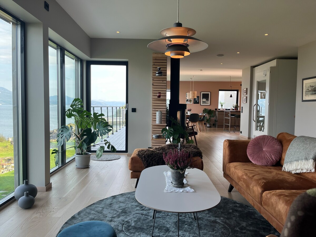 A spacious living area features large glass windows, providing views of the ocean and surrounding landscape. Green plants add life to the room, while a comfortable couch and a round table invite relaxation. Natural light fills the space, highlighting modern decor.