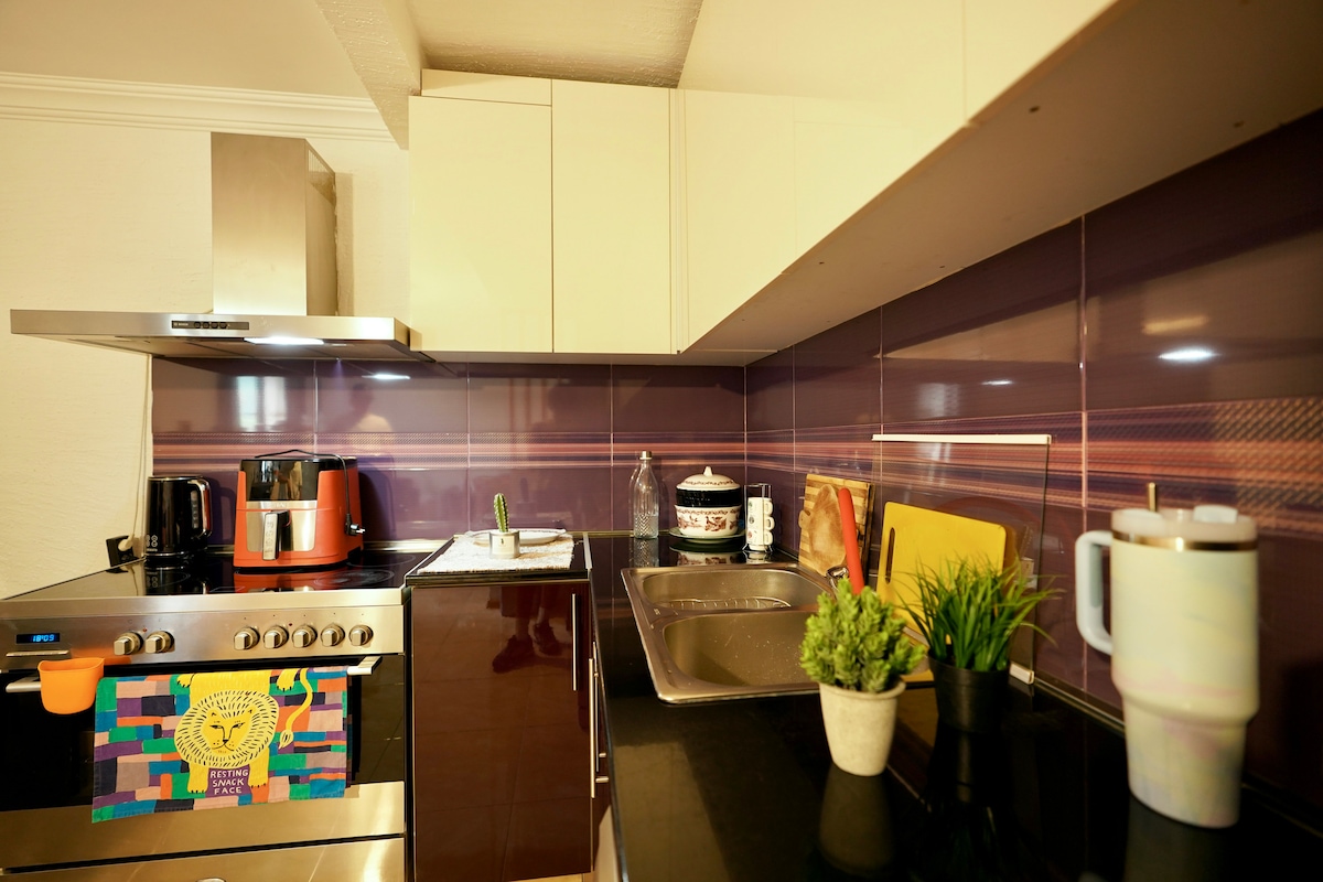 The kitchen features modern appliances including a stainless steel oven and a coffee maker. A sleek countertop holds a small potted plant and a decorative mug. Back-painted purple tiles add a contemporary touch to the space.