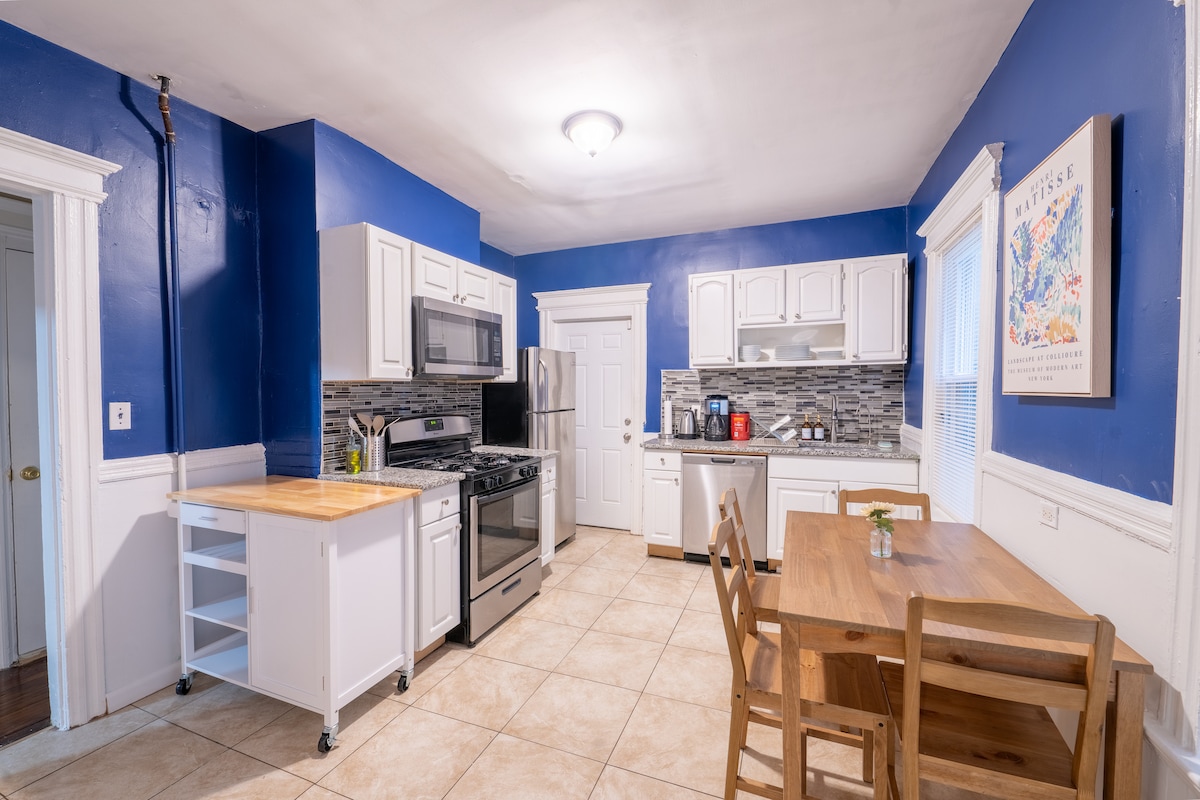 The kitchen features dark blue walls and white cabinetry with a wooden countertop island. A stainless steel stove and microwave are prominently displayed. A wooden dining table with four chairs is positioned near the window. Tile flooring extends throughout the space.