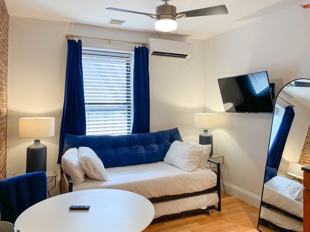A compact living area features a daybed with white bedding and two accent pillows. Nearby, a round white table sits between two blue armchairs. A large wall mirror reflects the space, while a mounted flat-screen TV and a ceiling fan add functionality. Bright light filters through the window with blue curtains.