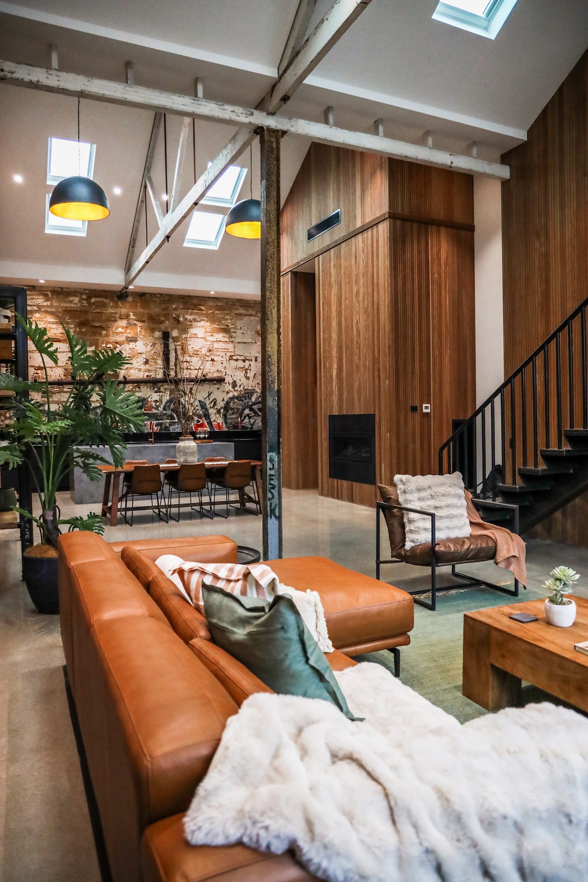 The spacious living area features a combination of natural wood and stone elements, with a comfortable leather sofa positioned prominently. A well-lit dining space is visible in the background, accompanied by large windows and a modern staircase leading to upper levels.