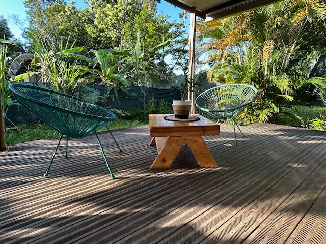 An outdoor terrace is furnished with two acapulco chairs and a wooden table, creating a relaxed seating area. Lush greenery surrounds the space, with various plants providing a natural backdrop. The wooden deck features a striped pattern that enhances the overall aesthetic.