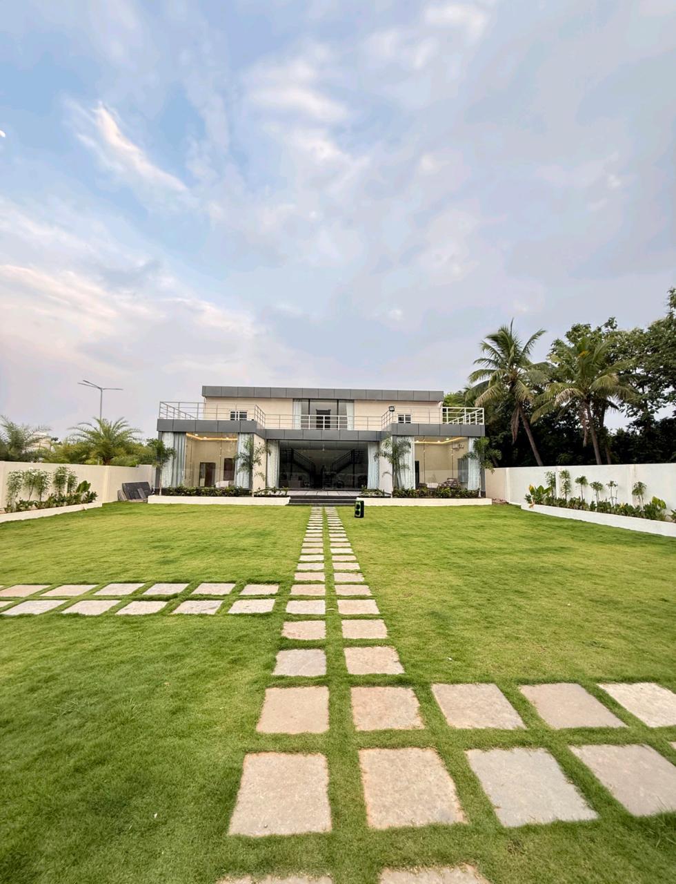 A modern two-story house is showcased against a partly cloudy sky. A manicured lawn with neatly arranged stepping stones leads to the entrance. Lush greenery and palm trees frame the property, contributing to an open and inviting atmosphere.