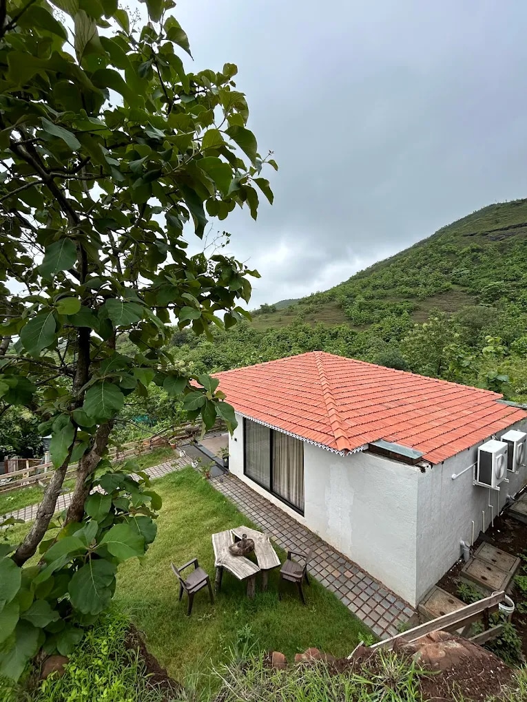 A cozy villa sits amidst lush greenery, featuring a terracotta roof and large glass doors that open to a tranquil outdoor area. A small wooden table and chairs are positioned on a stone patio, surrounded by grassy terrain and rolling hills in the background.