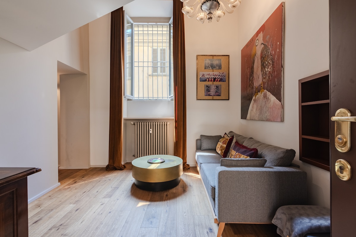 A contemporary living area is shown with a light wooden floor and a grey sofa adorned with decorative pillows. A round coffee table sits in the center, while a bright window with a grid design allows natural light to enter. Wall art adds character to the space.