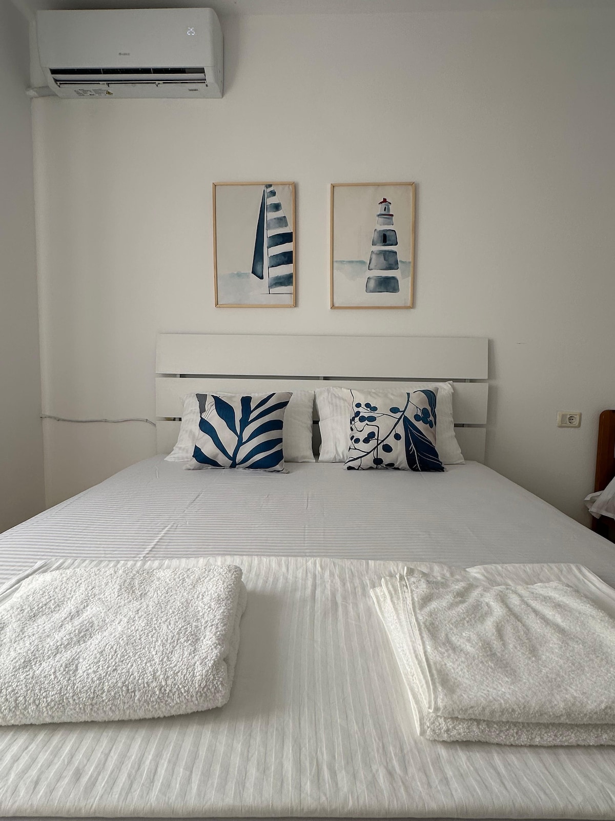 A spacious sleeping area is presented with a neatly made bed, featuring white linens and decorative pillows with nature-inspired patterns. Two framed artworks depicting lighthouses adorn the wall above the headboard, adding a subtle coastal touch to the room.