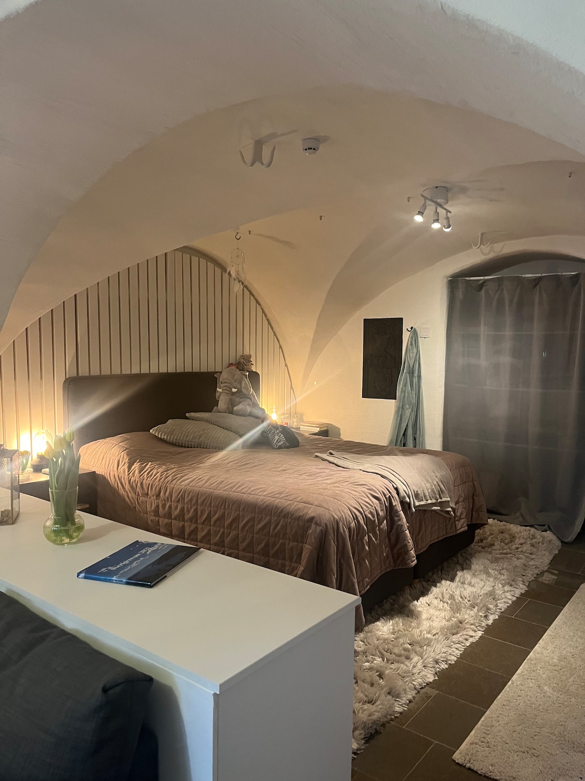 A spacious sleeping area features vaulted ceilings and soft lighting. A large bed is dressed with neutral linens, flanked by decorative pillows. A cozy rug lies beneath, and a small table holds a vase with flowers. Natural light filters in through a partially draped window.