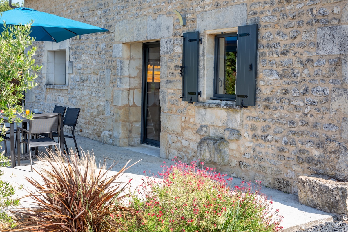 The exterior of the renovated barn displays natural stone walls with dark shutters and a door leading inside. A shaded outdoor seating area is available, surrounded by vibrant plants and flowers on a stone patio.