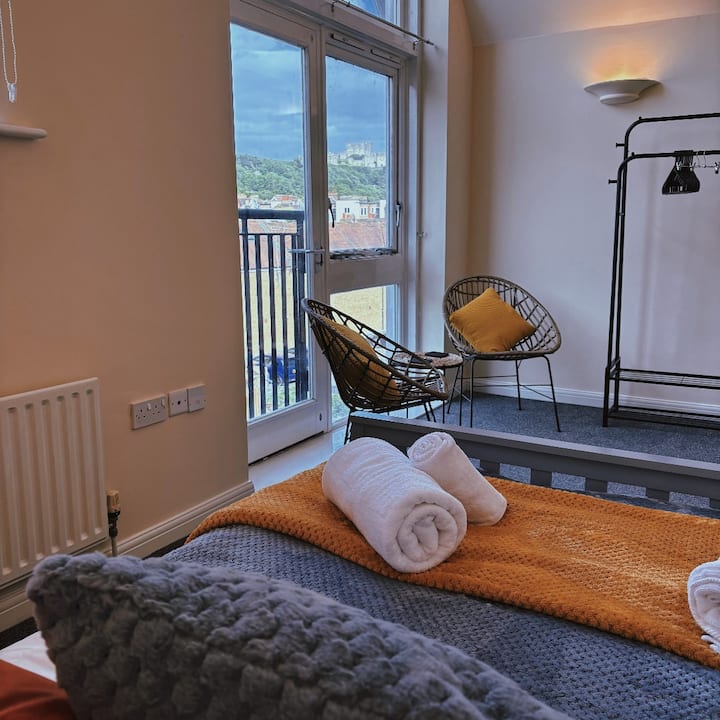 Este dormitorio luminoso cuenta con una cómoda cama con una colcha gris y naranja, dos toallas blancas cuidadosamente enrolladas a los pies y modernas sillas de mimbre con cojines amarillos. Una gran ventana se abre a una vista panorámica, añadiendo encanto y luz natural.