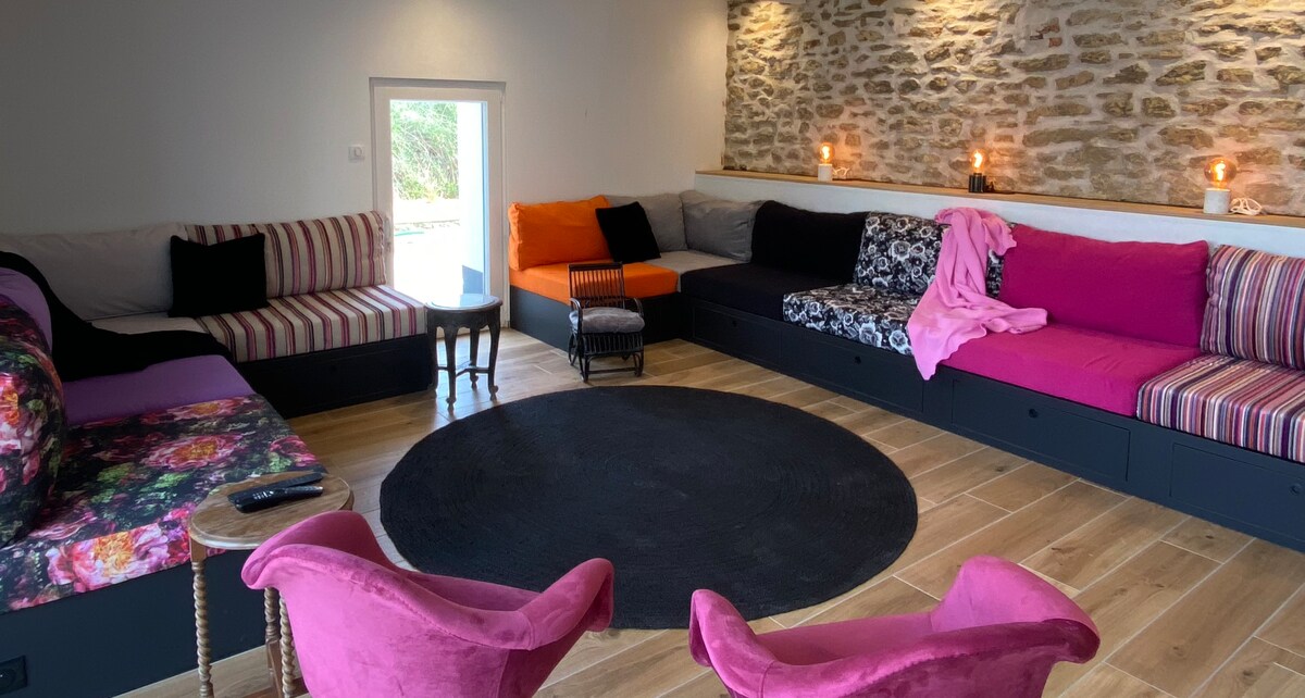 A spacious lounge area features multiple low seating arrangements in various colors and patterns, including stripes and floral designs. Soft lighting from wall sconces creates a warm ambiance, while a circular rug anchors the room. A corner table offers a small surface for drinks or decor.