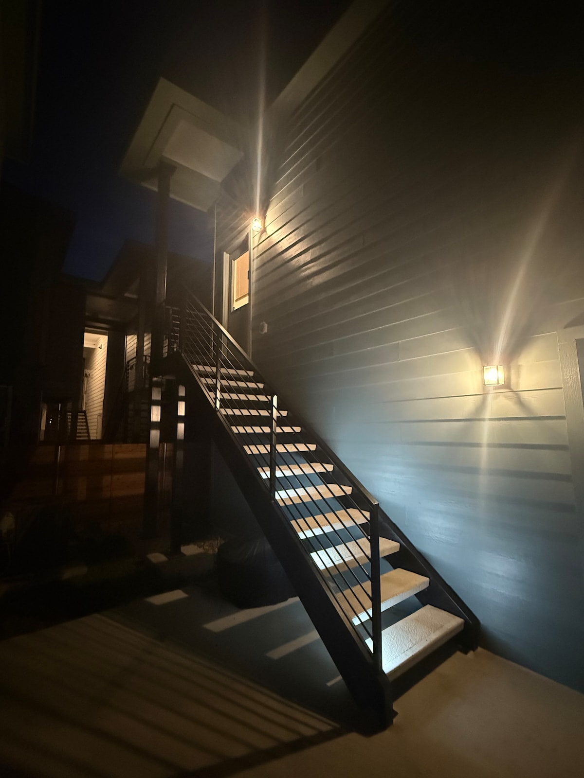 An exterior view of a modern building is captured at night, highlighting illuminated stairs leading up to a doorway. Soft lighting accentuates the staircase railings and walls, creating a welcoming approach to the entrance.