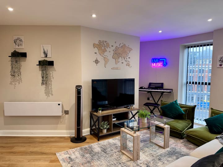 غرفة معيشة أنيقة بأرضيات خشبية وتلفزيون بشاشة مسطحة على حامل خشبي ومقاعد مبطنة باللون الأخضر. تشمل الديكورات نباتات معلقة، وخريطة العالم على الحائط، ولافتة "موسيقى حية" من النيون بجانب لوحة المفاتيح لإضفاء لمسة نابضة بالحياة.
