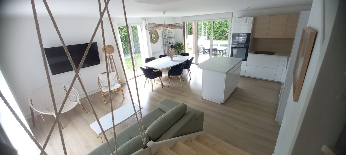 A spacious living area features a modern design with light wooden flooring and large glass doors leading to an outdoor space. A white kitchen island offers a functional space, complemented by a dining table surrounded by dark chairs. A green sofa provides comfort and relaxation.