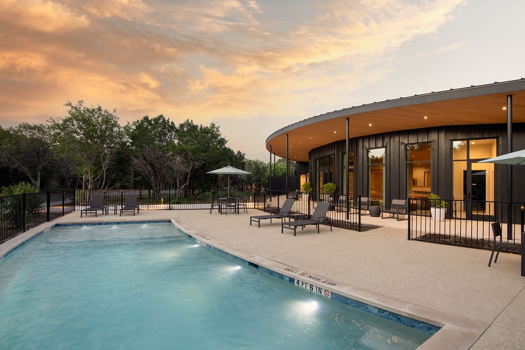 A curved deck surrounds a serene swimming pool, set against a backdrop of trees and a sunset sky. Comfortable lounge chairs are positioned near the pool, with umbrellas providing shade. Large windows of the building reflect the warm hues of dusk, enhancing the peaceful atmosphere.