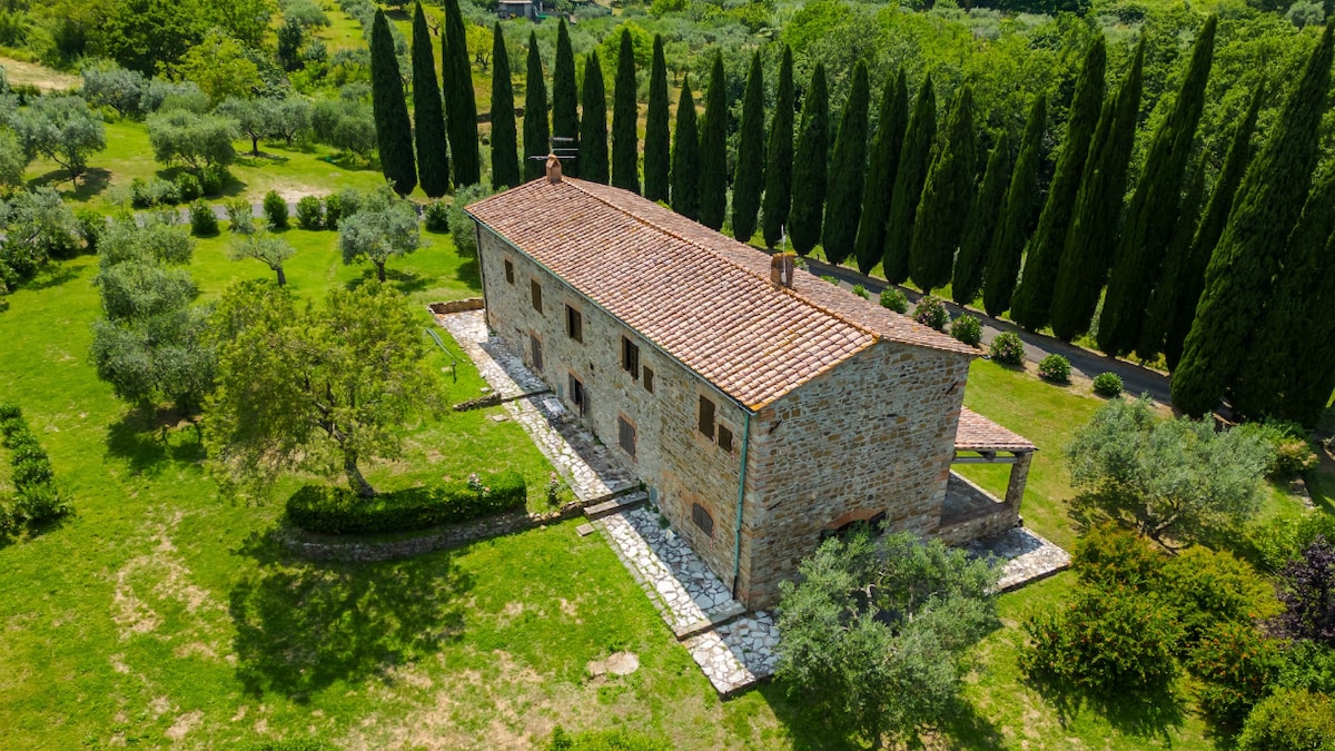 An aerial view captures a rustic stone farmhouse nestled among lush greenery. Olive trees and tall cypress trees surround the property, highlighting its tranquil rural setting. A stone pathway leads to the entrance, emphasizing the home's connection to the natural landscape.