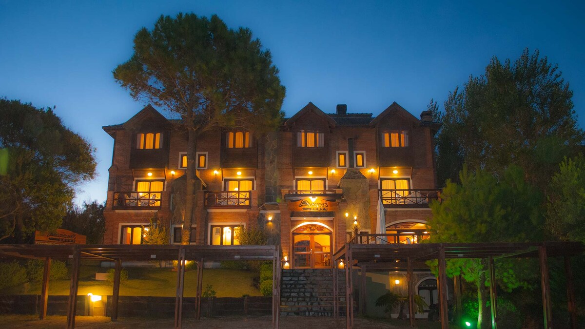 The exterior of a three-story building is illuminated, showcasing a warm ambiance against a twilight backdrop. The structure features multiple balconies and windows, framed by surrounding trees and a well-maintained garden space, enhancing the welcoming appearance of the space.