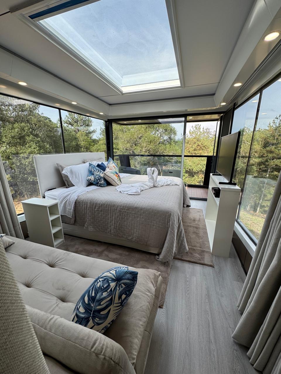 The stylish bedroom features large glass panels offering scenic views of the surrounding greenery. A spacious bed with soft linens is complemented by decorative pillows. A cozy seating area is present, alongside a flat-screen TV mounted on the wall. Natural light brightens the space through the skylight.