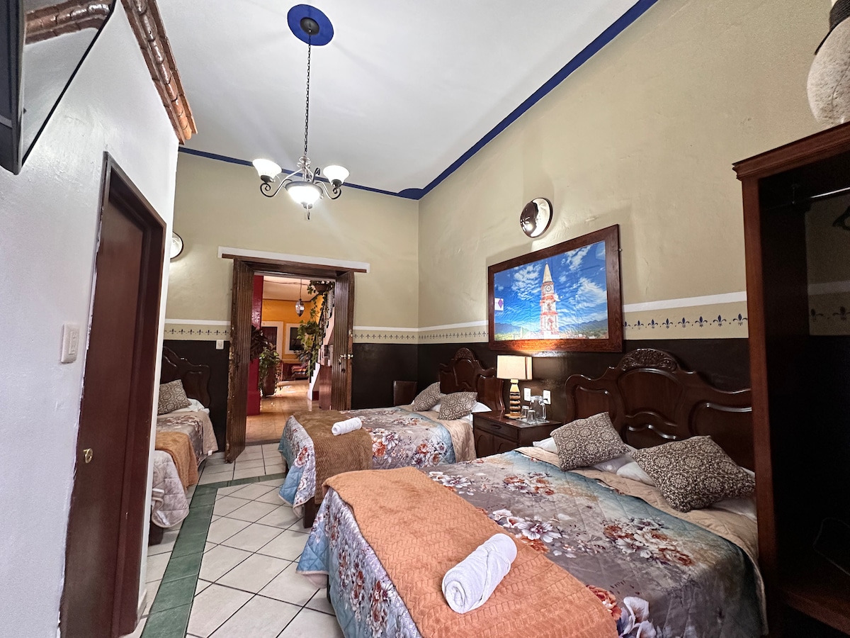 A spacious room features two matrimonial beds and one individual bed, each adorned with floral bedding. A large television is mounted on the wall, and soft lighting is provided by decorative fixtures. The tile floor extends throughout, with a glimpse of the doorway leading to another area visible.