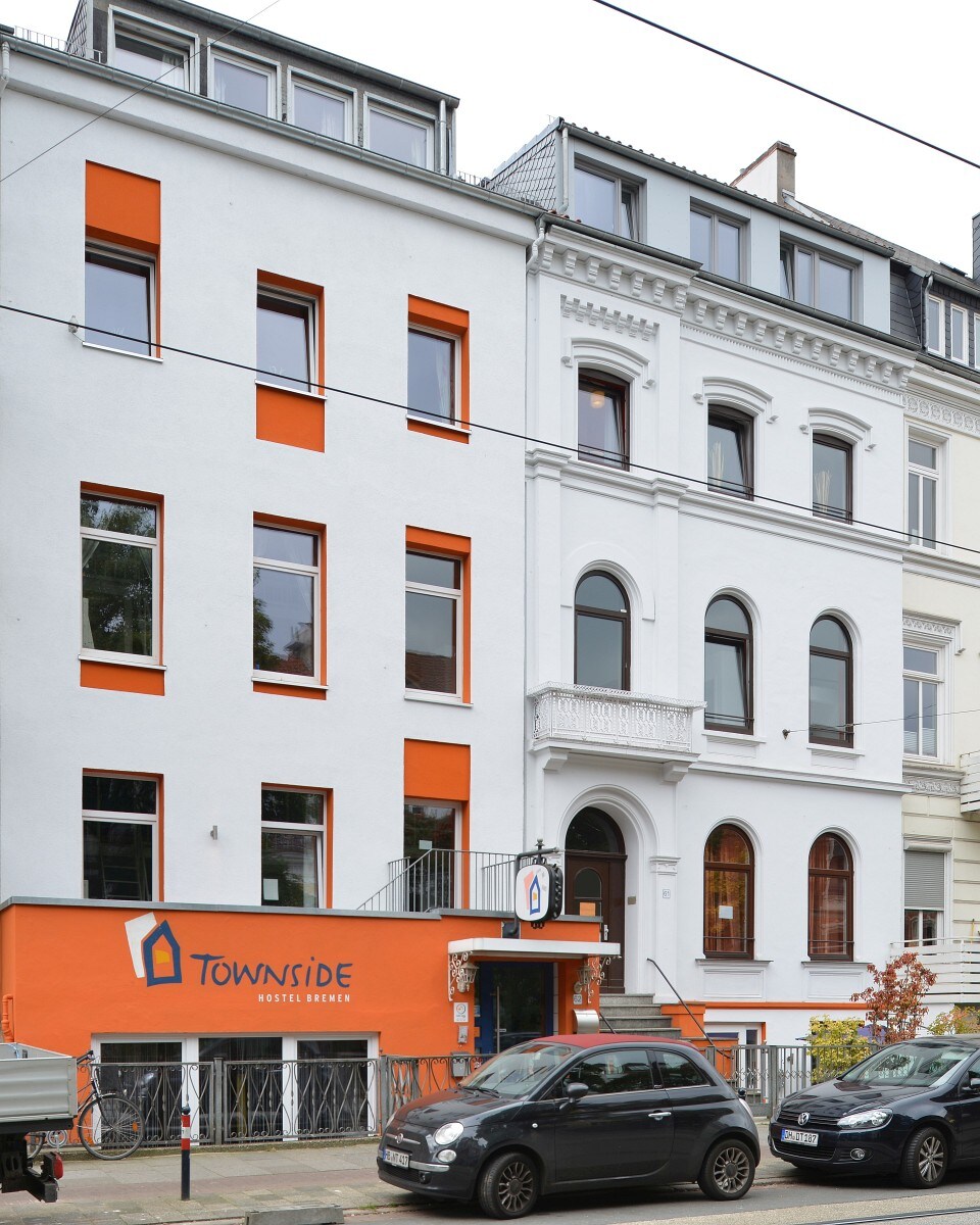 The exterior of the hostel is depicted, showcasing a white building with vibrant orange accents. Large windows provide natural light, and a welcoming entrance is visible. Nearby vehicles are parked along the street, contributing to a lively urban atmosphere.