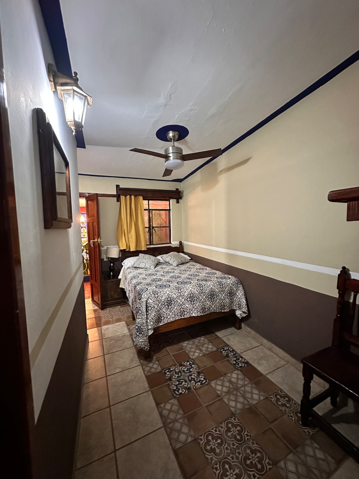 A narrow hallway leads to a cozy bedroom featuring a double bed with a blue and white patterned coverlet. A ceiling fan is present above, and a window with yellow curtains allows natural light into the space. Traditional tile flooring adds texture to the room.