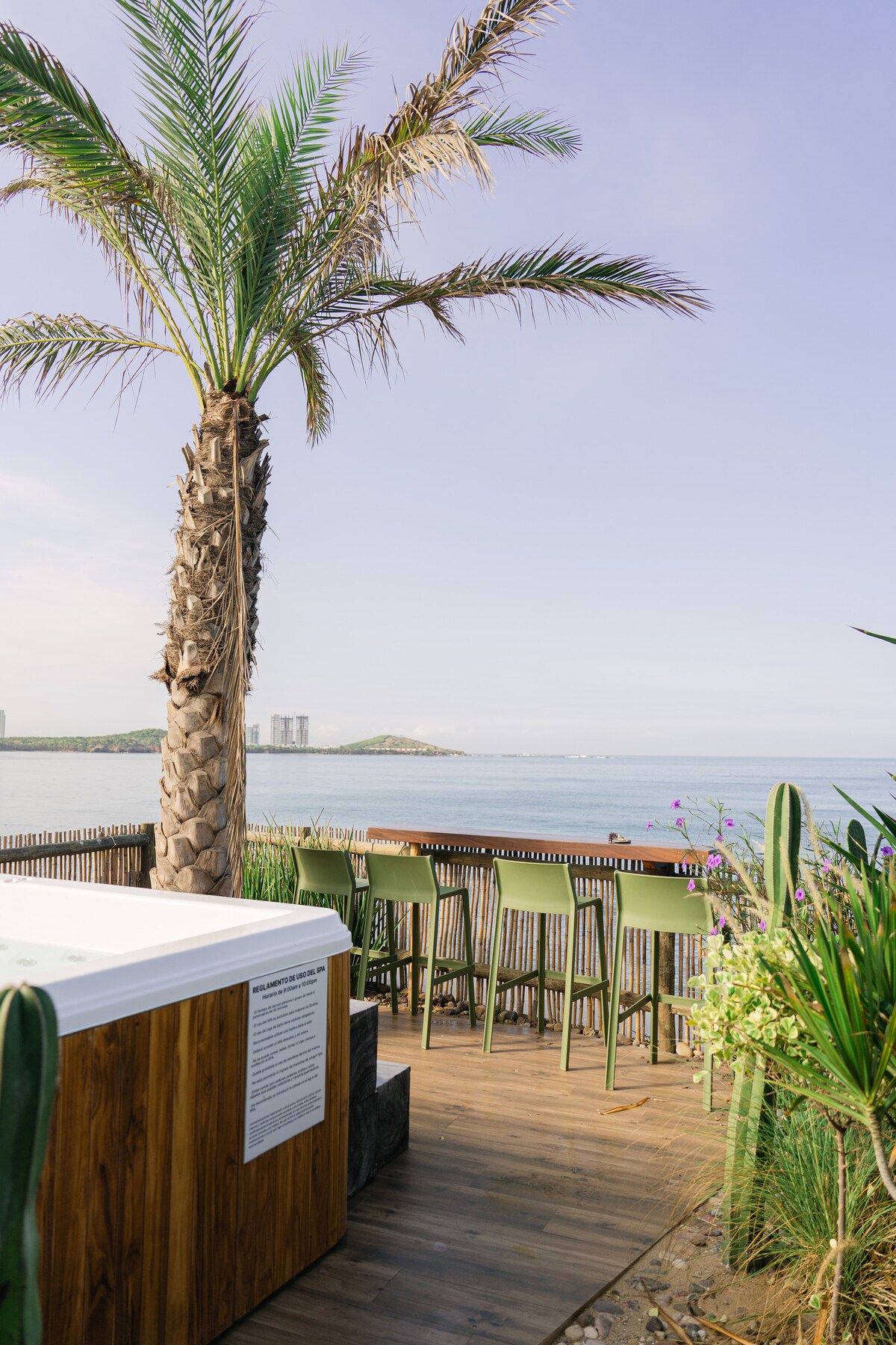 A scenic outdoor area features a hot tub surrounded by lush greenery and a palm tree. Bar seating is present, offering a view of the calm sea and distant land. Natural light enhances the tranquil atmosphere, making it an ideal spot for relaxation.